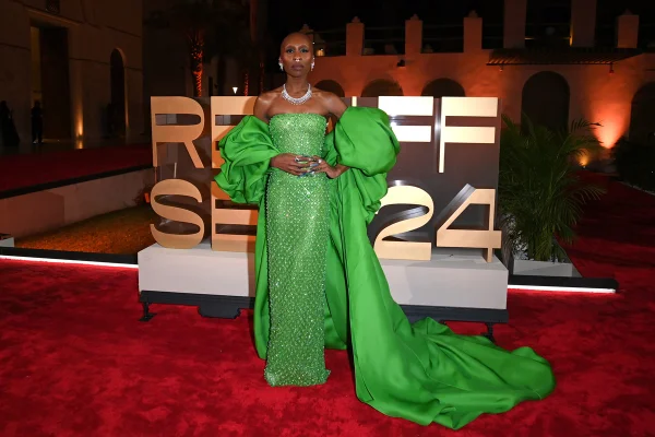 A imagem mostra uma mulher negra posando em um tapete vermelho. Com as mãos na cintura, ela olha para a câmera usando um vestido verde longo