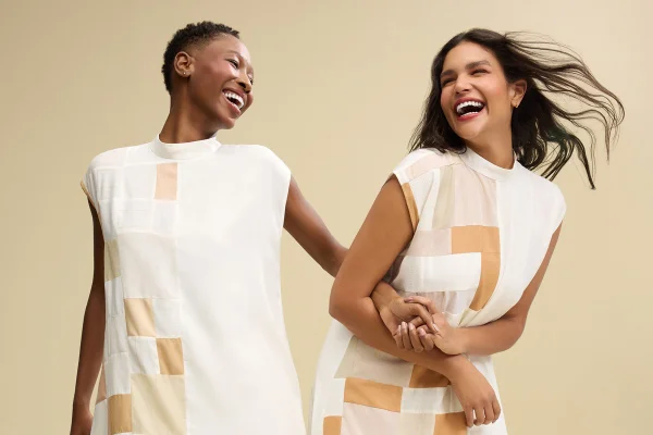 Duas mulheres de braços dados e rindo. Elas usam roupas brancas - Metrópoles
