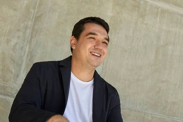 Na foto, vemos um homem de camiseta branca e blazer preto olhando para longe e sorrindo