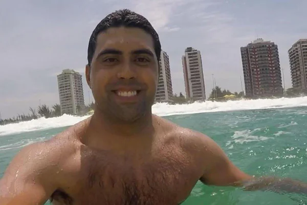 foto colorida do rosto e ombros de homem dentro de mar com cidade ao fundo - metrópoles