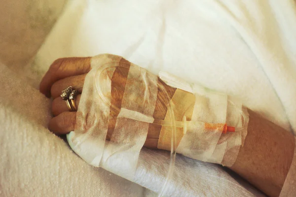 Foto colorida de mão feminina, idosa, com curativos e tubos hospitalares. Metrópoles.