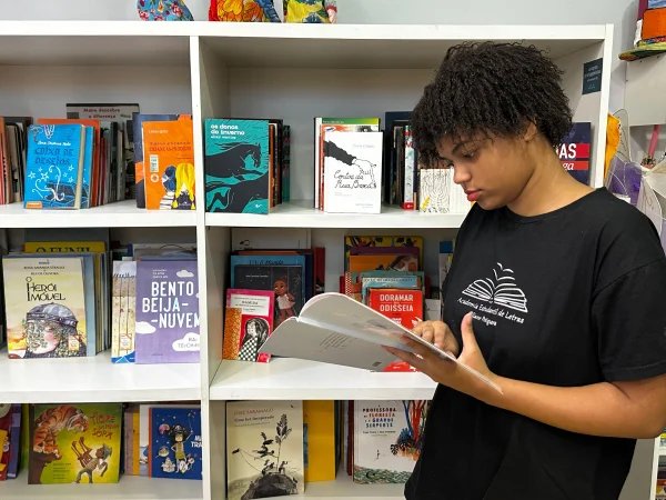 Fotografia colorida mostra estudante lendo livro em biblioteca-Metrópoles