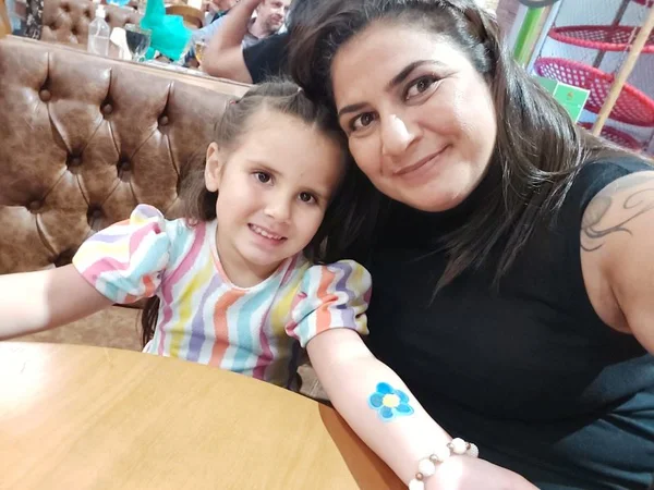 imagem colorida mostra mulher ao lado de criança em foto. a mulher é branca, tem cabelo liso e veste uma blusa preta. a criança veste uma blusa colorida. metrópoles