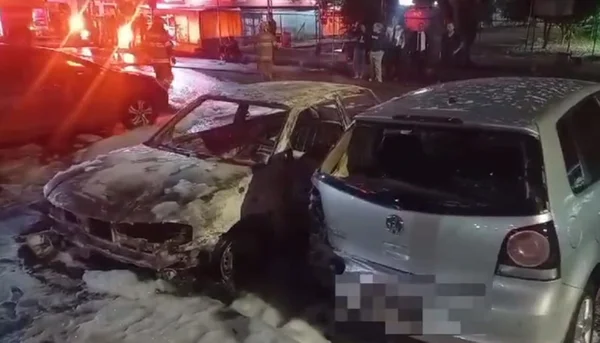 Dois carros em estacionamento. Carro completamente queimado à esquerda e carro prata levemente queimado à direita.