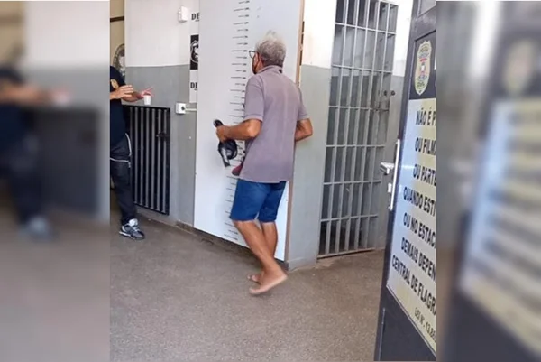 foto de um homem idoso caminhando de costas em uma delegacia. ele está com as pés descalços e veste uma bermuda azul e uma camisa cinza
