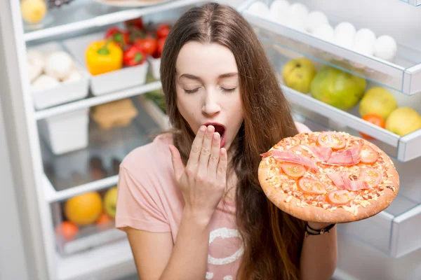 Foto colorida de mulher bocejando em frente a uma geladeira e segurando uma pizza - Metrópoles