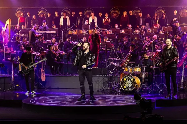 foto colorida de banda de quatro integrantes em palco com orquestra no fundo - metrópoles