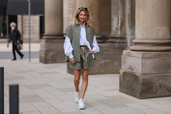 Mulher branca e de meia idade, com cabelo ondulado loiro, andando pelas ruas de Paris. Ela veste uma camisa de botão branca, um shorts e um colete bege e um tênis branco