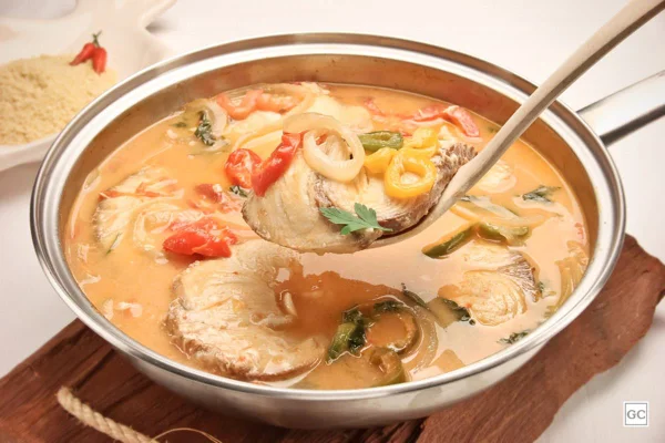 Peixe cozido no leite de coco em frigideira de metal. Uma concha segura um pedaço de peixe - Metrópoles