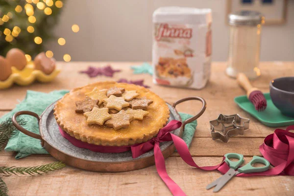 Na foto vemos uma mesa de madeira com uma torta decorada com estrelas e luzes de natal ao fundo