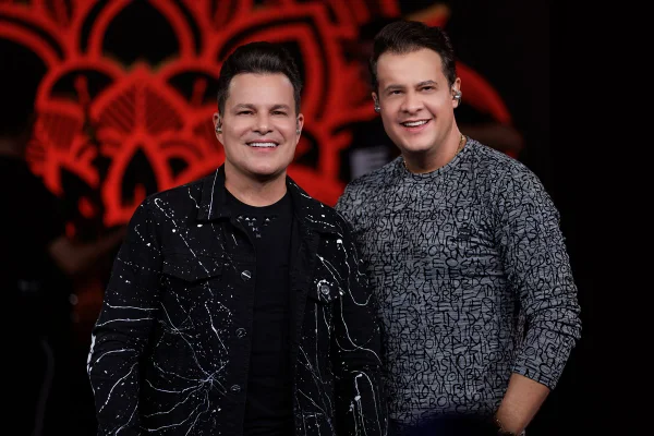 foto de dois homens brancos, um está de camiseta preta e jaquela preta com desenhos brancos e o outro com uma blusa de manga longa granulada em preto e branco