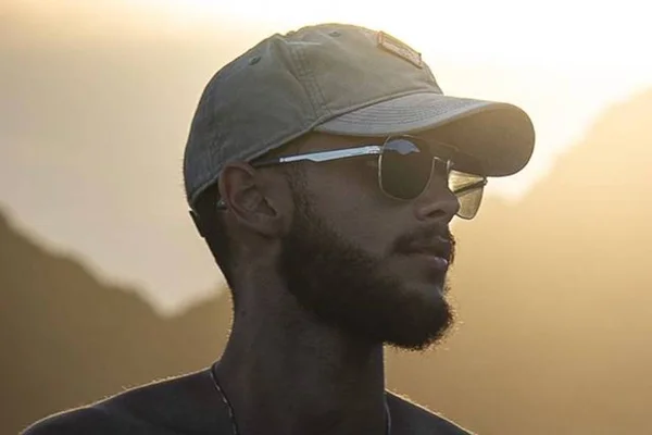 Foto colorida de Guilherme Rocha, irmão de Chay Suede, de bon[é e óculos escuros no pôr do sol - Metrópoles