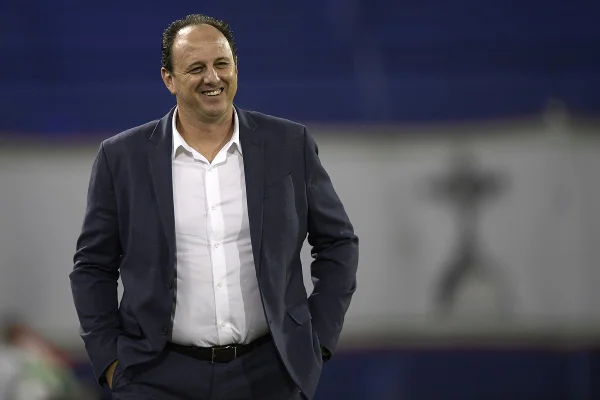 Imagem colorida de homem vestido de terno com sorriso aberto em fundo branco com azul - Metrópoles