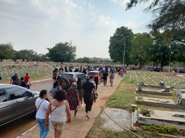 Imagem de pessoas acompanhando um velório no cemitério de Taguatinga. Ao redor da capela, há flores e itens em tons verdes
