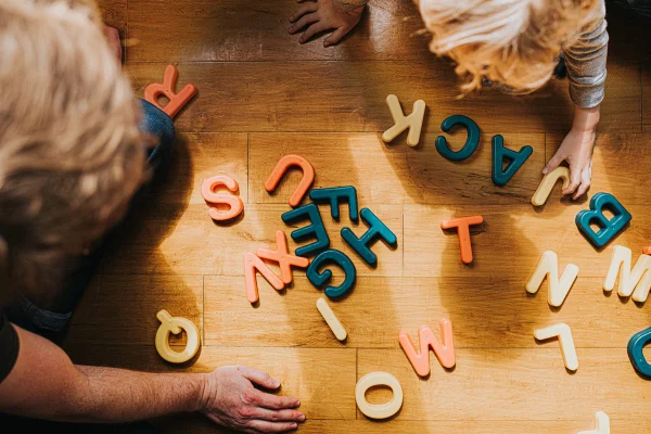 A criança organiza letras maiúsculas coloridas para criar palavras. Os pais supervisionam e ajudam.