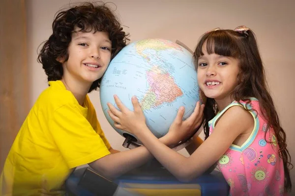 um menino branco de cabelo cacheado e blusa amarela segura um globo terrestre de frente a ele também com o globo m mãos uma menina branca de cabelos lisos castanhos e vestido rosa. Os dois seguram o globo e sorriem