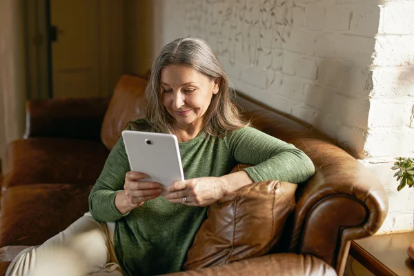 Pessoas, envelhecimento e dispositivos eletrônicos: mulher de meia-idade com longos cabelos grisalhos relaxando em casa, usando um tablet para se comunicar online com a família e sorrindo ao olhar para a tela. Metrópoles