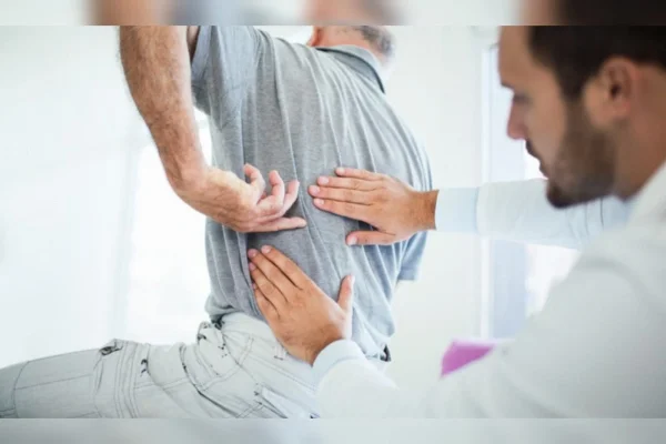 Um idoso sentado na maca apontando para a coluna e o médico examinando o local