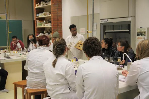 Imagem colorida de cientista sentados em volta de uma mesa; um homem negro está de pé, ao centro, mostrando um cilindro transparente - Metrópoles