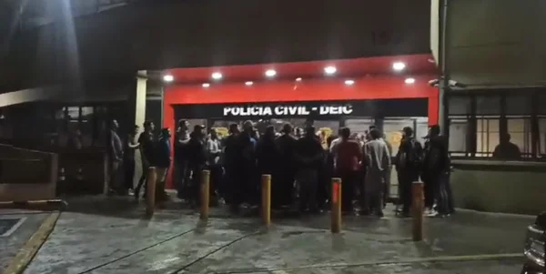 Imagem de porta de delegacia com pessoas reunidas; elas foram vítimas de um golpe feito por pai e filho - Metrópoles