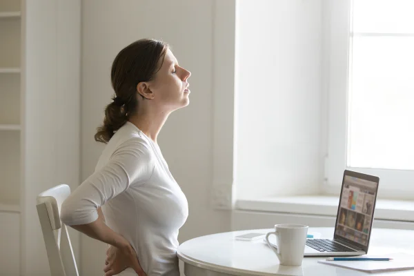Imagem mostra mulher de roupas claras sentada em uma mesa e alongando as costas - Metrópoles
