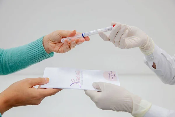Imagem colorida de mulher entregando tubo de exame para profissional de Saúde - Metrópoles