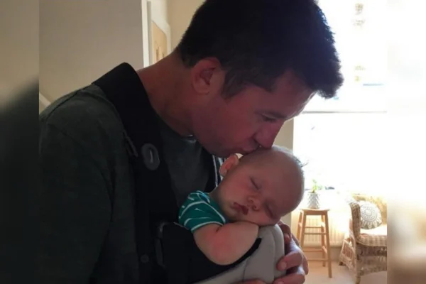 Foto colorida e um pouco escura de um homem beijando um bebê na cabeça, enquanto a criança dorme - Metrópoles.