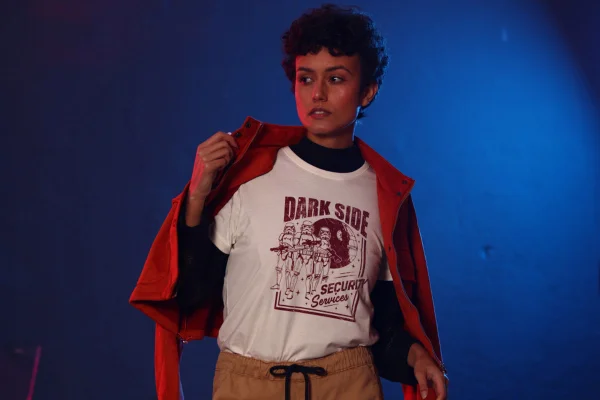 Mulher morena, jovem e com cabelo cacheado curto, posando para foto em um fundo escuro. Ela usa uma camiseta branca com estampa do filme Star Wars, um casaco vermelho e uma calça marrom clara. - Metrópoles