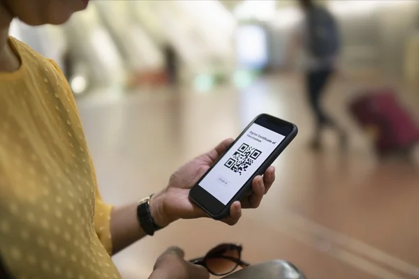 Mulher branca mexendo no celular. Ela usa um relógico com correia preta de couro e uma blusa amarela. Na tela do aparelho é possível ver um QR Code