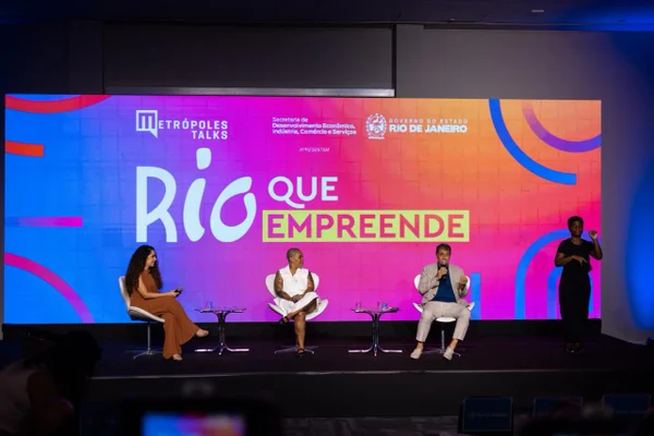Visão mais aberta do palco, com três pessoas sentadas e uma mediadora. O telão ao fundo exibe a identidade visual “Rio que Empreende”, em letras grandes e coloridas. A plateia aparece em sombra, assistindo ao debate
