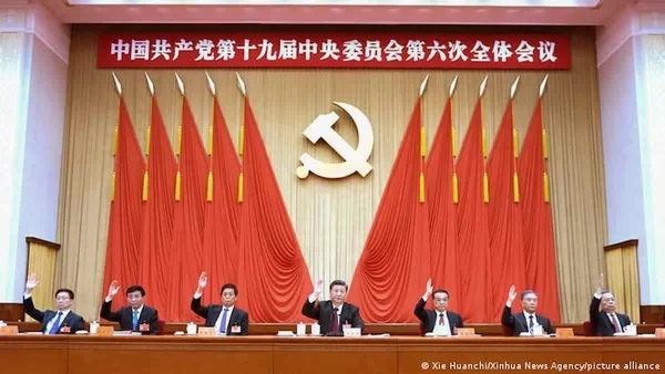Salão principal da sede do Partido Comunista Chinês. Sete homens, incluindo o presidente chinês Xi Jin Ping, erguem as mãos em votação, e ao fundo é possível ver bandeiras vermelhas, escritos em ideogramas e o símbolo do comunismo - Metrópoles