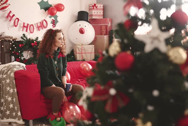 Imagem colorida: sala com decoração de Natal e mulher sentada em sofá olhando para o lado - Metrópoles