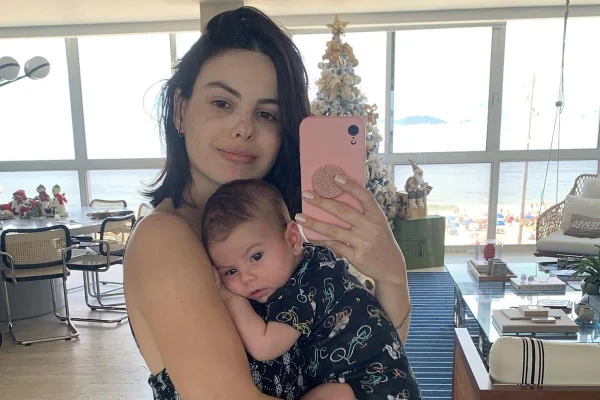 A atriz Sthefany Brito, uma mulher branca com cabelo liso e castanho, posando para foto em frente ao espelho, com o celular, e o filho no colo.
