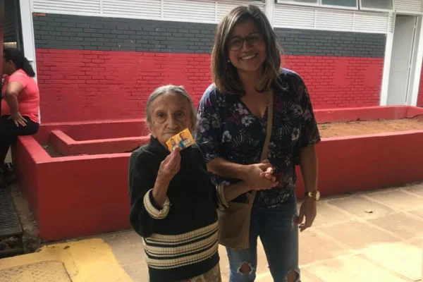 Foto colorida de mulher jovem segurando a mão de idosa, enquanto sorri para foto. -Metrópoles.