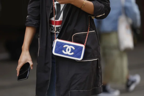 Pessoa andando pela rua com camiseta branca, casaco preto e uma bolsa transpassada da marca Chanel. O item é pequeno e retangular, na cor azul, com a logo da marca na cor branca