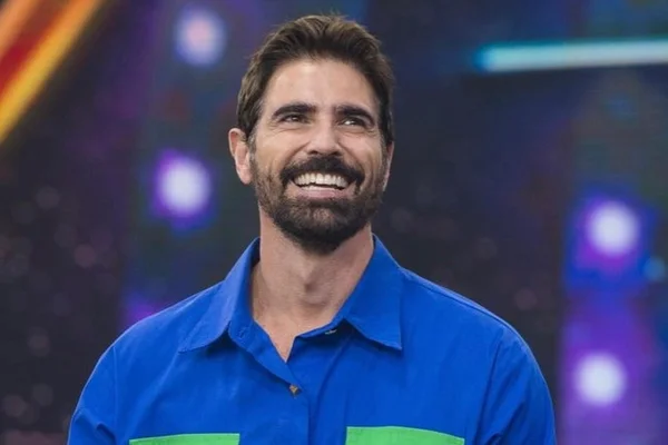 Reynaldo Gianecchini de camiseta polo azul sorrindo em palco de programa - metrópoles