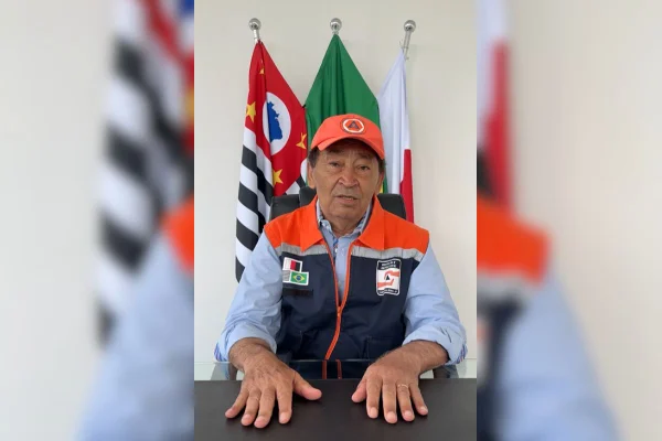 Imagens coloridas mostram homem sentado à mesa, de colete azul e laranja, boné laranja, sentado atrás de mesa, ao fundo bandeiras de São Paulo (à esquerda), do Brasil (centro) e de Taboão da Serra (à direita)