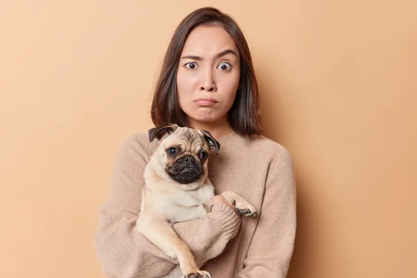 Foto colorida de mulher com expressão facial incomum, segurando um cachorro em seus braços - Metrópoles.