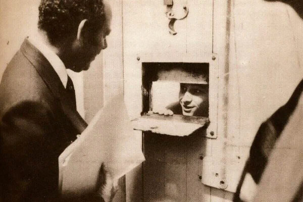 Imagem em preto e branco de preso em bocal de porta de aço falando com homem engravatado - Metrópoles-
