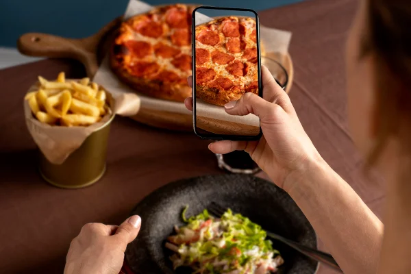 Foto colorida de mulher segurando celular, enquanto fotografa com o aparelho uma pizza, mas consome comida saudável. - Metrópoles.