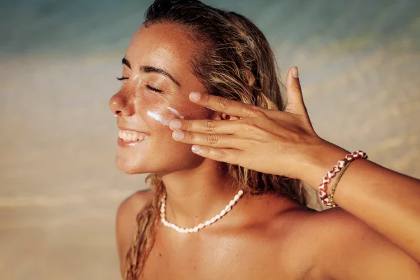 Foto colorida de mulher na praia e aplicando protetor solar no rosto - Metrópoles