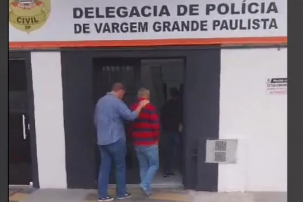 Homem com camisa polo com grossas listras horizontais, é conduzido, de costas, por policial civil à entrada de delegacia onde há a inscrição "delegacia de polícia de vargem grande paulista" - Metrópoles
