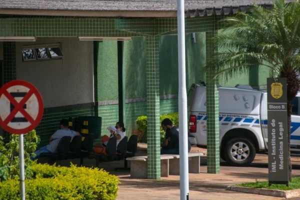 Pessoas sentadas em bancos na frente do IML; carro estacionado