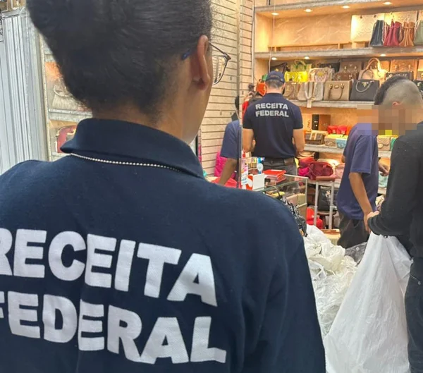 Agente da Receita Federal olha para comércio; pessoas seguram sacolas grandes - Metrópoles