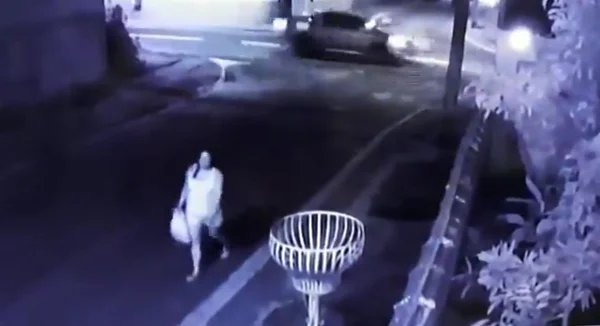 Mulher caminha na rua e, ao fundo, um carro passa em alta velocidade