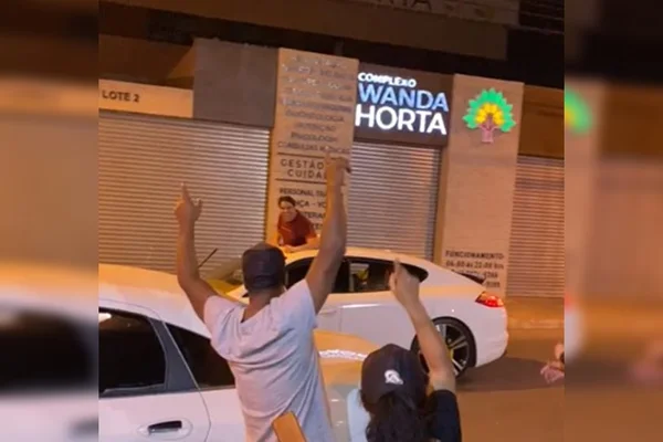 Homem em teto solar de carro branco e pessoas de costas com a mão para cima, à noite