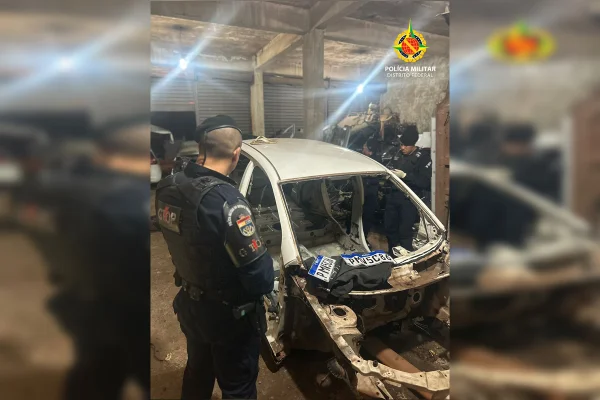 Imagem colorida de um carro sem a maioria das peças. À esquerda, um policial militar de farda azul observa o carro