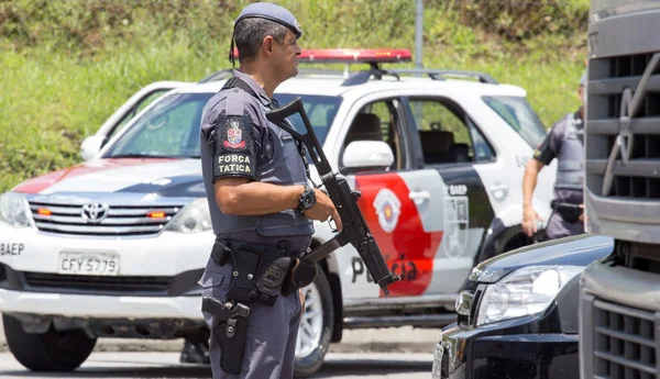 Foto colorida de policial militar armado com viatura da PM ao fundo - Metrópoles