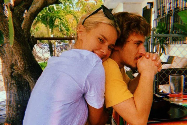 Na imagem colorida, mulher de blusa branca abraça rapaz de roupa amarela. Os dois estão sentados em uma mesa, em um jardim - Metrópoles