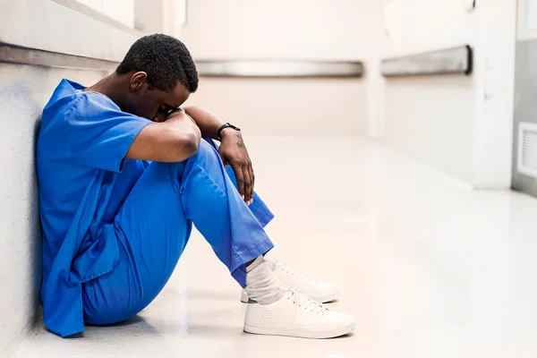 Médico sentado no chão, de corpo inteiro. Profissional médico cansado no corredor. Ele está de uniforme azul no hospital. - Metrópoles